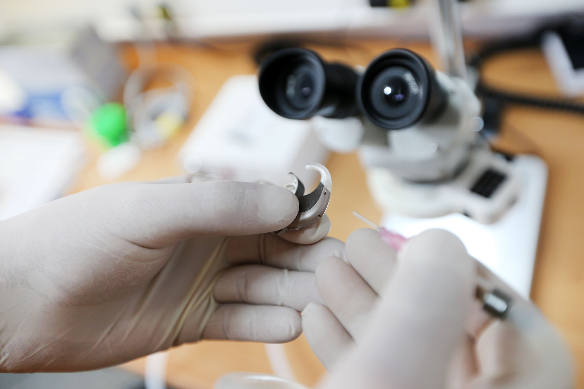 Hände mit Gummihandschuhen reparieren ein Hörgerät. Mikroskop im Hintergrund.